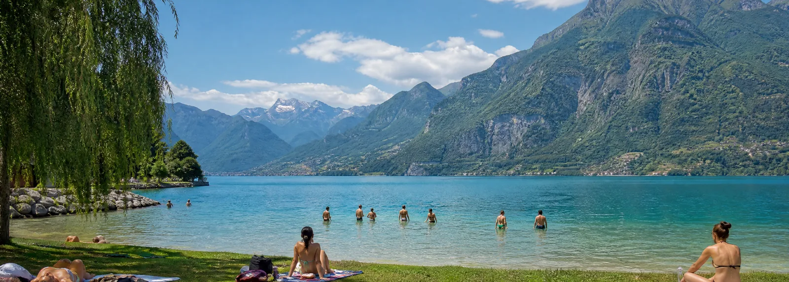 A grassy lakeside beach with swimmers in shallow water and mountains on the far shore