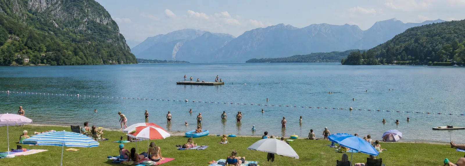 Bagnanti, un pontile per i tuffi e file di boe su una spiaggia lacustre erbosa d'estate